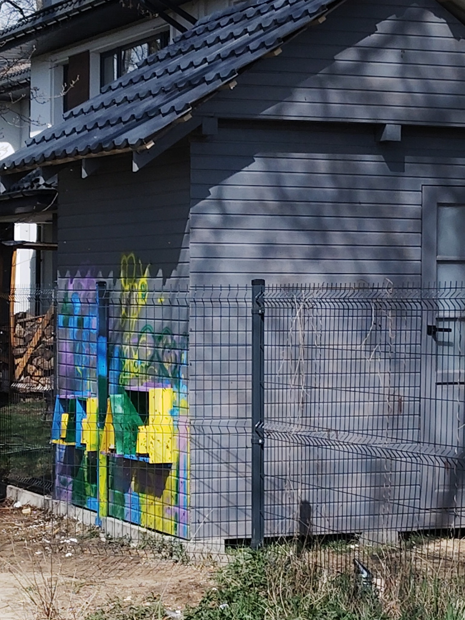 Szara drewniana altana z ciemnym dachem, z kolorowymi graffiti na bocznej ścianie, widoczna przez metalową siatkę ogrodzeniową.