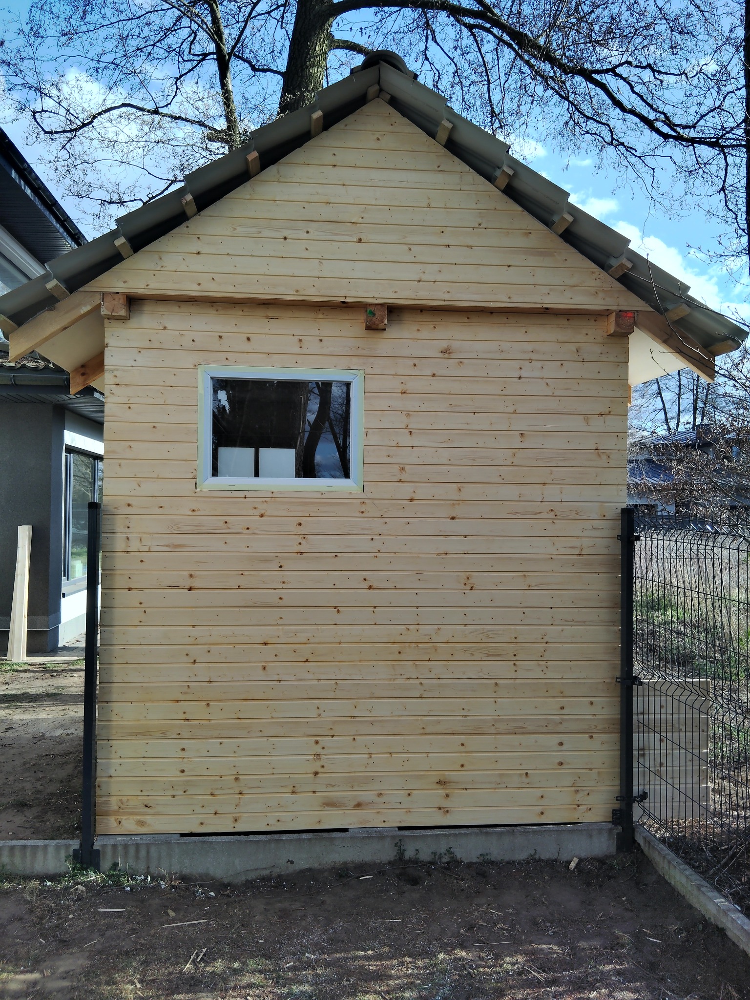 Nowo wybudowany, surowy drewniany domek narzędziowy z oknem, zadaszony szarą blachą, widoczny z boku, na tle błękitnego nieba i drzew bez liści, ogrodzony metalową siatką.