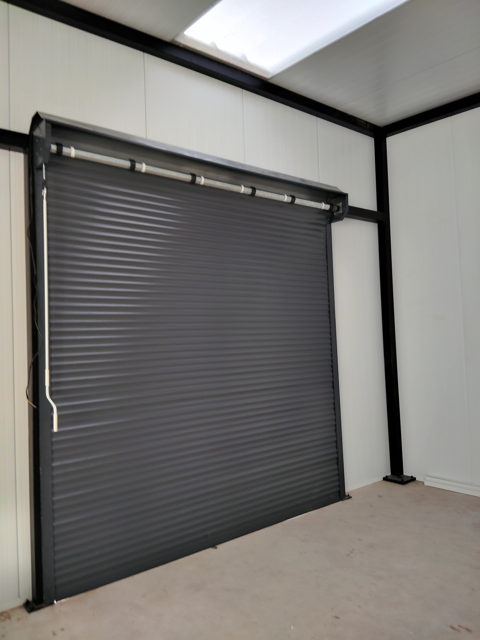 Zamknięta, szara brama garażowa rolowana z czarną ramą w jasnym pomieszczeniu z białymi ścianami panelowymi i betonową podłogą.
