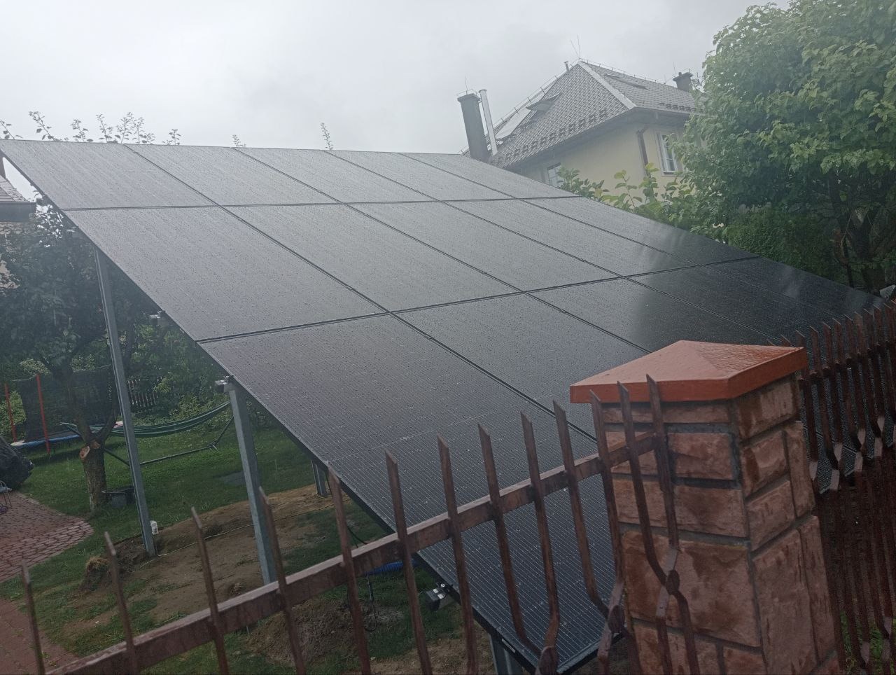 Panel fotowoltaiczny na stelażu, widoczny zza metalowego ogrodzenia w deszczowy dzień, z domem w tle.