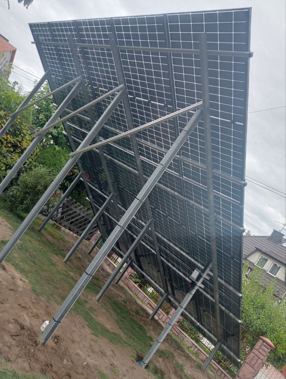 Widok od tyłu na panele fotowoltaiczne zamontowane na metalowej konstrukcji na działce, z widoczną instalacją i elementami mocującymi, w tle dom jednorodzinny i zieleń.