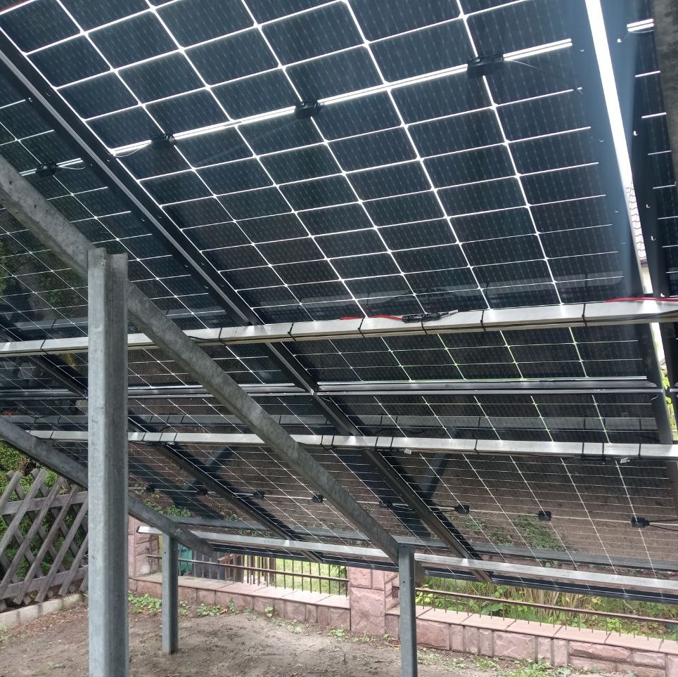 Widok od spodu na panele słoneczne zamontowane na metalowej konstrukcji, z widocznymi przewodami i ogrodzeniem w tle.