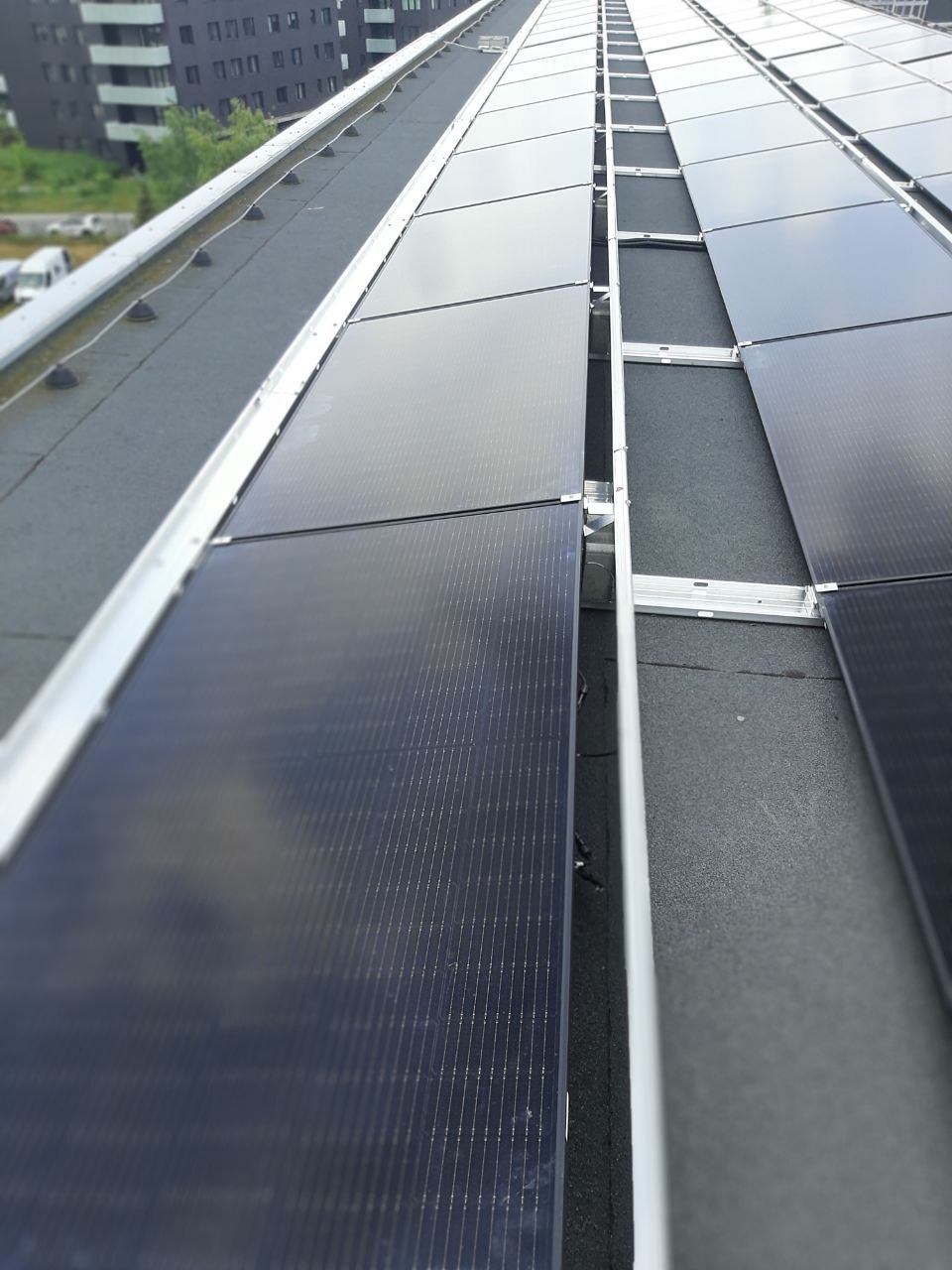 Rząd paneli fotowoltaicznych zamontowanych na płaskim dachu budynku, widok z góry pod kątem, z widocznymi elementami konstrukcyjnymi i budynkiem w tle.