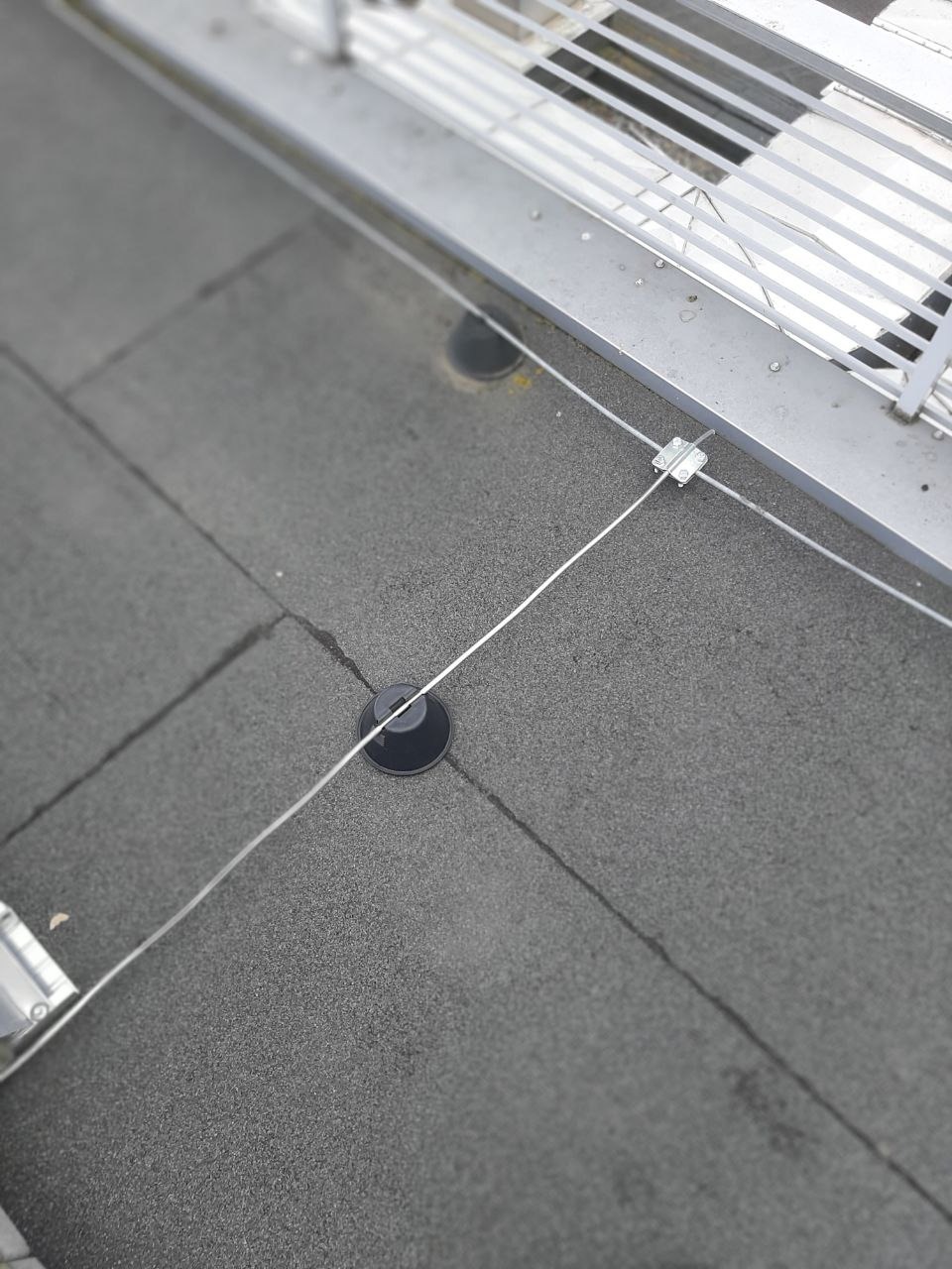 Szary dach pokryty papą z zamontowanymi uchwytami do odgromu i linką odgromową, widok z góry z lekkim rozmyciem.