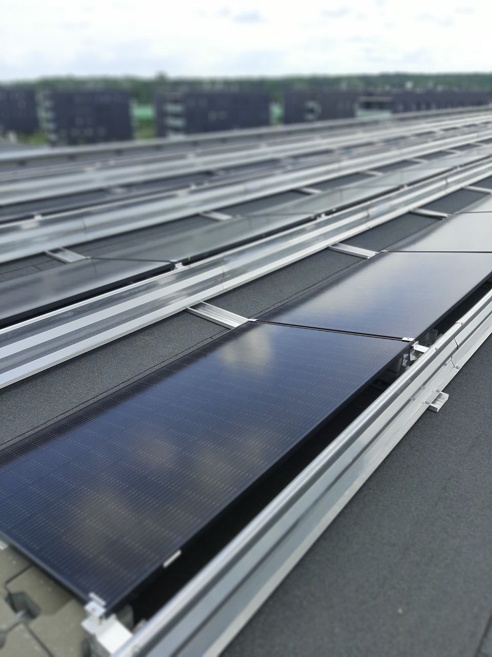 Rząd paneli fotowoltaicznych zamontowanych na płaskim dachu budynku, widoczne aluminiowe konstrukcje wsporcze i czarna powierzchnia dachu, budynki w tle lekko rozmyte.