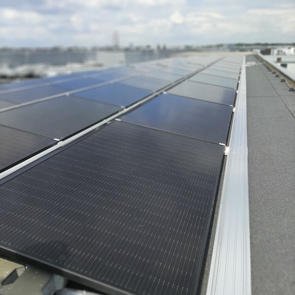 Rząd paneli fotowoltaicznych na płaskim dachu, widok z boku, z widoczną konstrukcją montażową i szarą membraną dachową.