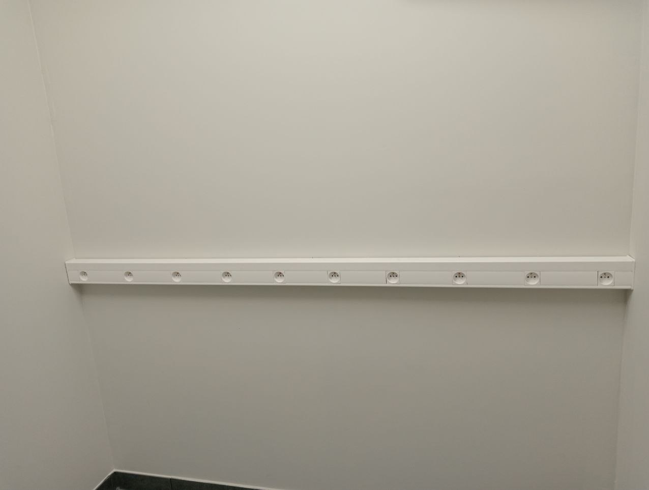 Biała listwa z gniazdkami elektrycznymi zamontowana poziomo na białej ścianie w rogu pomieszczenia, widoczne czarne panele podłogowe.