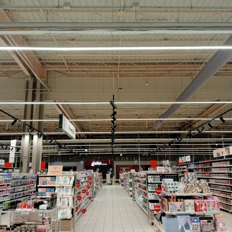 Wnętrze sklepu z oświetleniem liniowym LED podwieszonym do sufitu, widok na alejki z regałami pełnymi towarów.