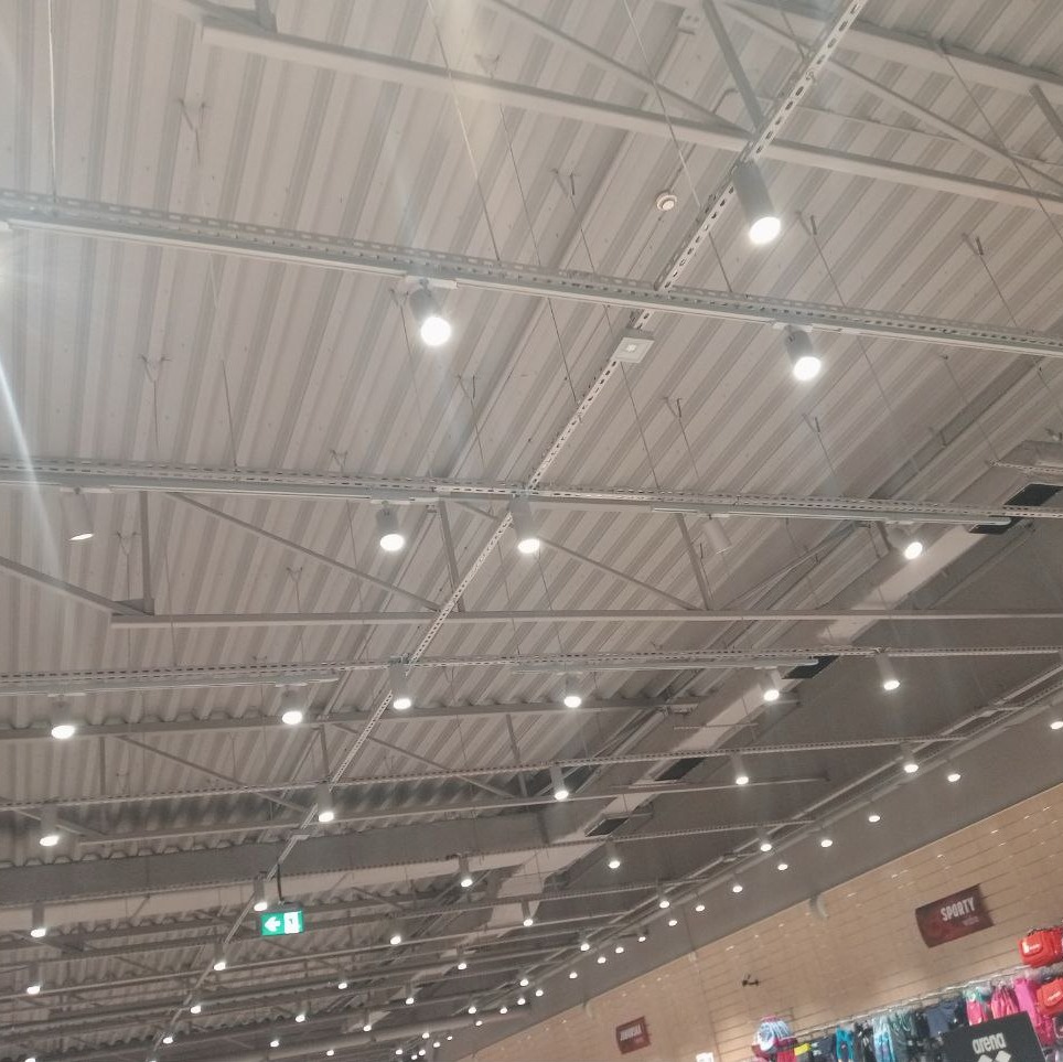 Widok sufitu w sklepie z zamontowanymi szynami i punktowym oświetleniem LED, widoczne elementy instalacji elektrycznej i system wentylacji.