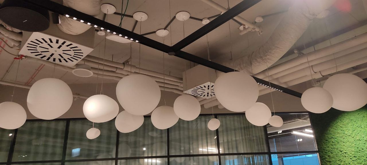 Nowoczesne oświetlenie biurowe: białe, kuliste lampy wiszące na tle sufitu z widocznymi instalacjami i kratkami wentylacyjnymi, z fragmentem ściany z mchu po prawej stronie.