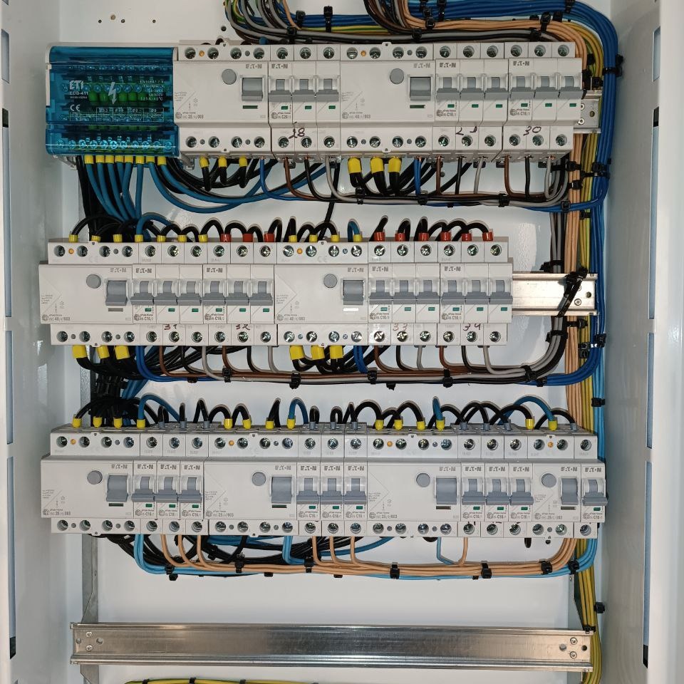 Rozdzielnica elektryczna z bezpiecznikami EATON, widoczne okablowanie w kolorach niebieskim, czarnym, żółto-zielonym i brązowym, zamontowana w białej szafce.