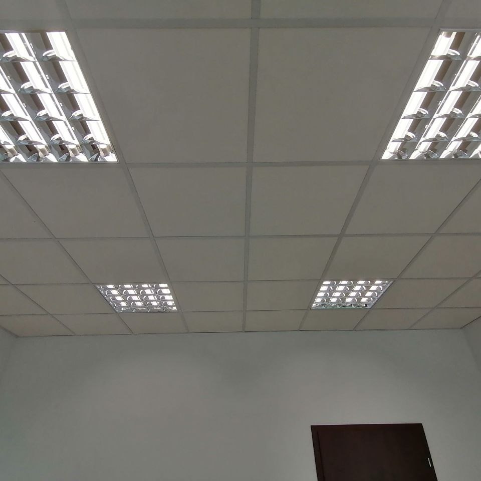 Kwadratowe panele sufitowe z rastrowymi oprawami oświetleniowymi, widok z dołu, fragment ściany z drzwiami w tle.