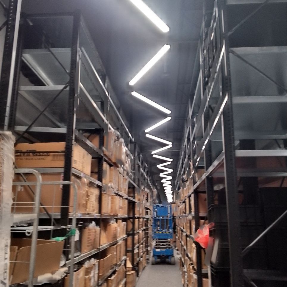 Długi korytarz magazynowy w Ząbkach z metalowymi regałami wypełnionymi kartonami, oświetlony rzędem lamp jarzeniowych i z podnośnikiem koszowym w oddali.