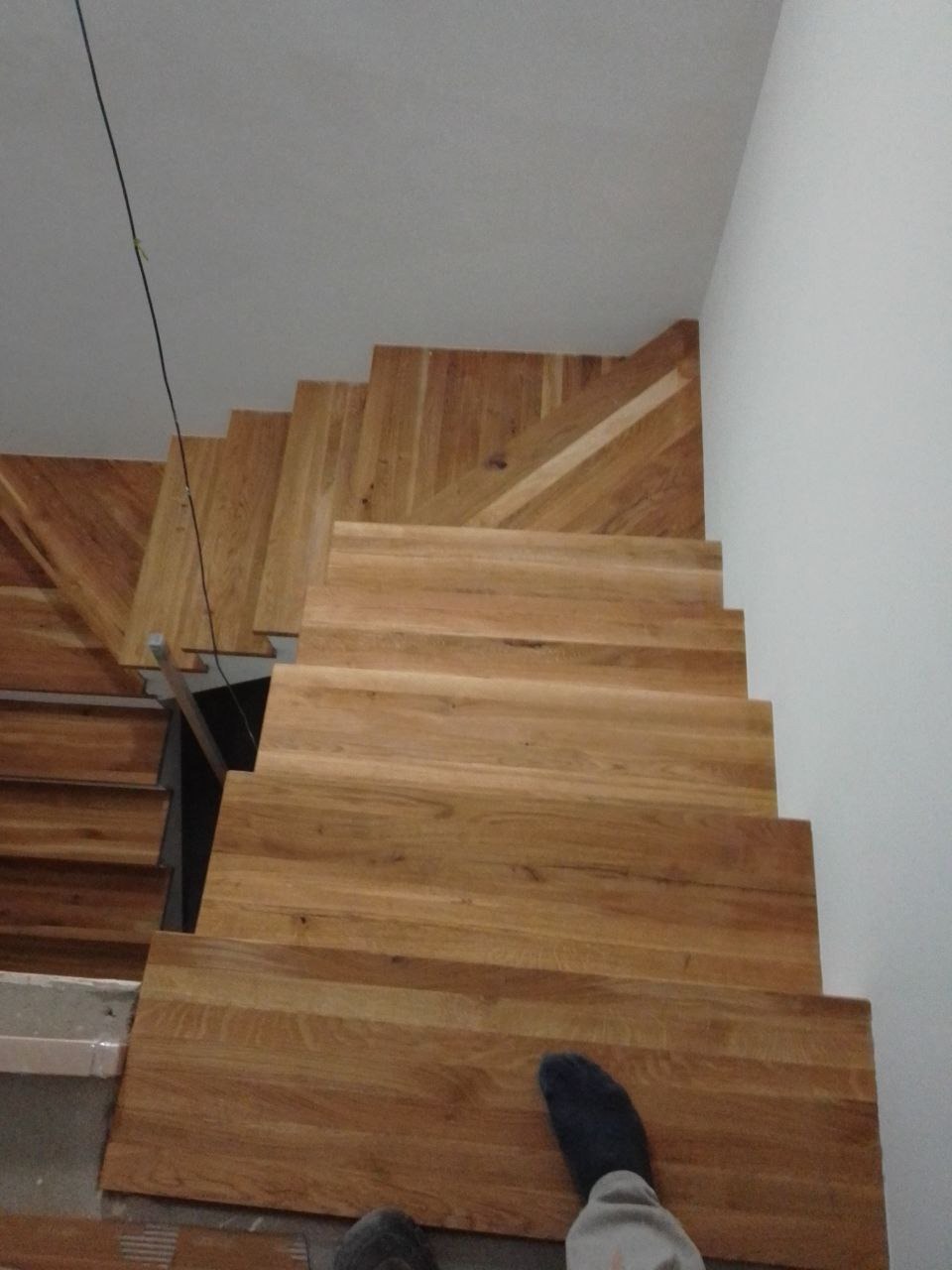 Montaż drewnianych schodów z widocznym fragmentem stopy w skarpetce, jasne drewno, surowy beton, częściowo ukończona instalacja.