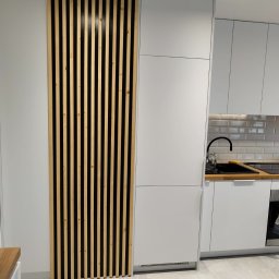 Drewniana Pszczółka Usługi Stolarskie Mateusz Pasieka - Biała kuchnia z drewnianą ścianką działową z pionowych listew, minimalistyczny design, jasne oświetlenie, szare płytki podłogowe. Nowoczesne wykończenie.