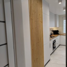 Drewniana Pszczółka Usługi Stolarskie Mateusz Pasieka - Biała zabudowa kuchenna z drewnianymi lamelami zdobiącymi front szafki, widok z perspektywy wnętrza, minimalistyczny design, jasne oświetlenie.