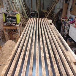 Drewniana Pszczółka Usługi Stolarskie Mateusz Pasieka - Równoległe, opalane deski z widocznym usłojeniem, ułożone na stole w warsztacie, z narzędziami i stosem drewna w tle. Drewno przygotowane do montażu.