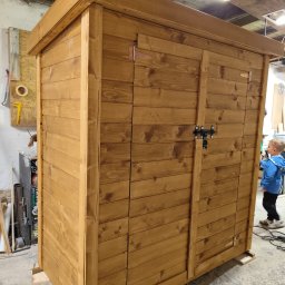 Drewniana Pszczółka Usługi Stolarskie Mateusz Pasieka - Drewniana, nowa, dwudrzwiowa szopa ogrodowa z metalowym zamknięciem, świeżo po impregnacji, stoi w warsztacie na podkładkach. W tle narzędzia i dziecko.