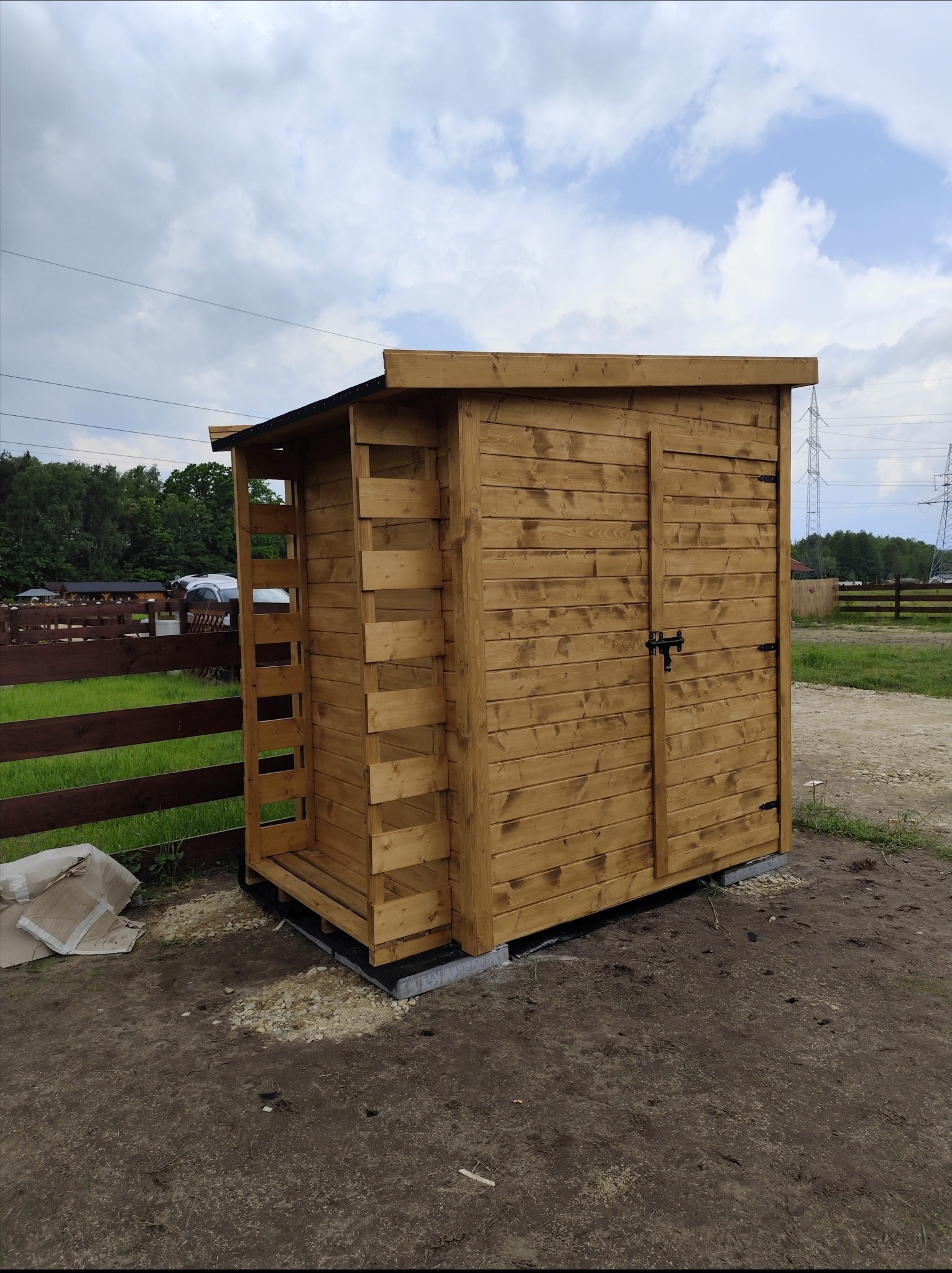 Drewniana, zamykana szopa ogrodowa z półkami, wykonana z jasnego drewna, z widoczną konstrukcją i zamykanymi drzwiami, na zewnątrz, w tle ogrodzenie i słupy wysokiego napięcia.