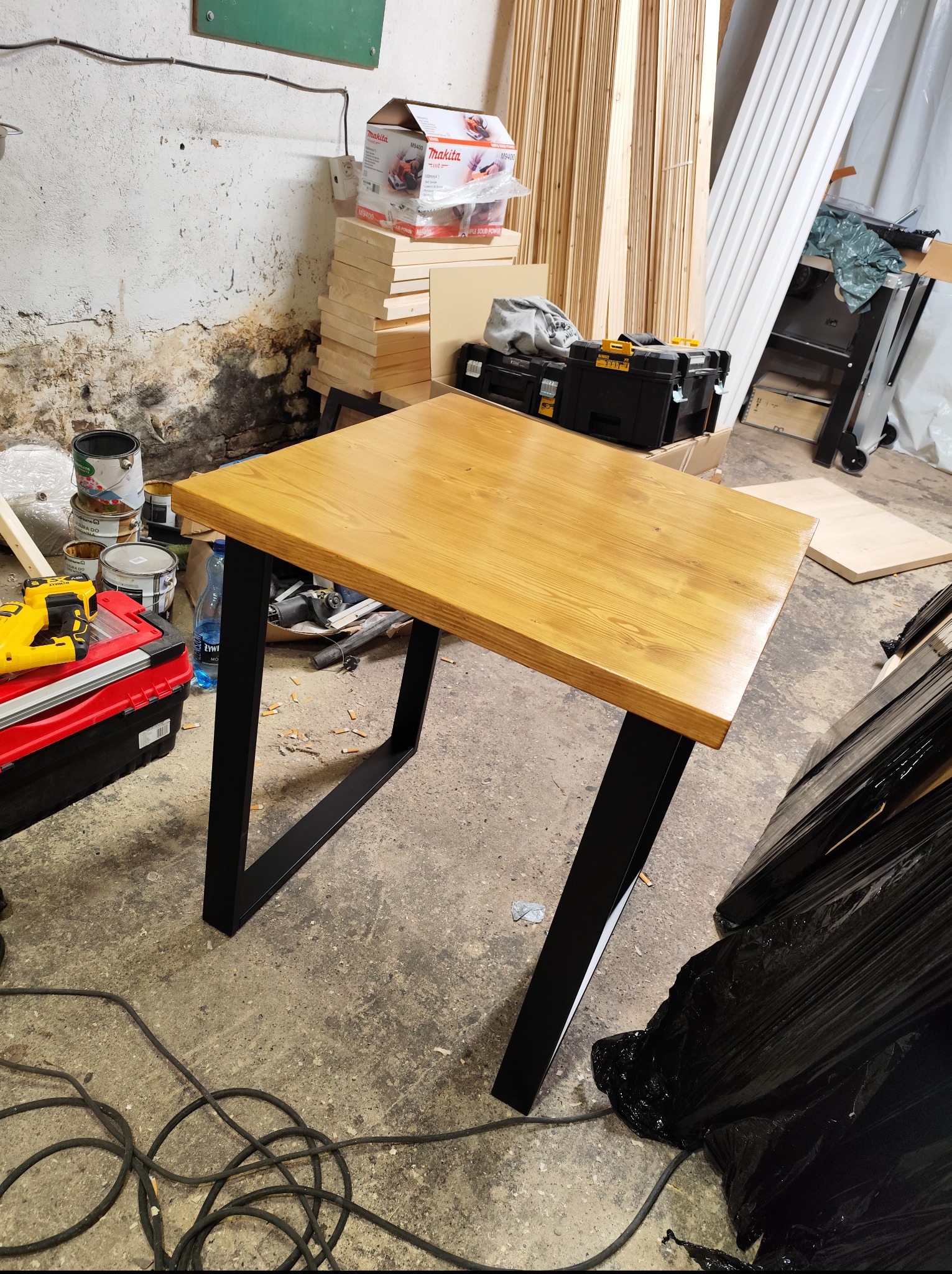 Stół z blatem z jasnego drewna i czarnymi, metalowymi nogami w kształcie litery V, stojący w warsztacie na tle narzędzi i materiałów budowlanych.