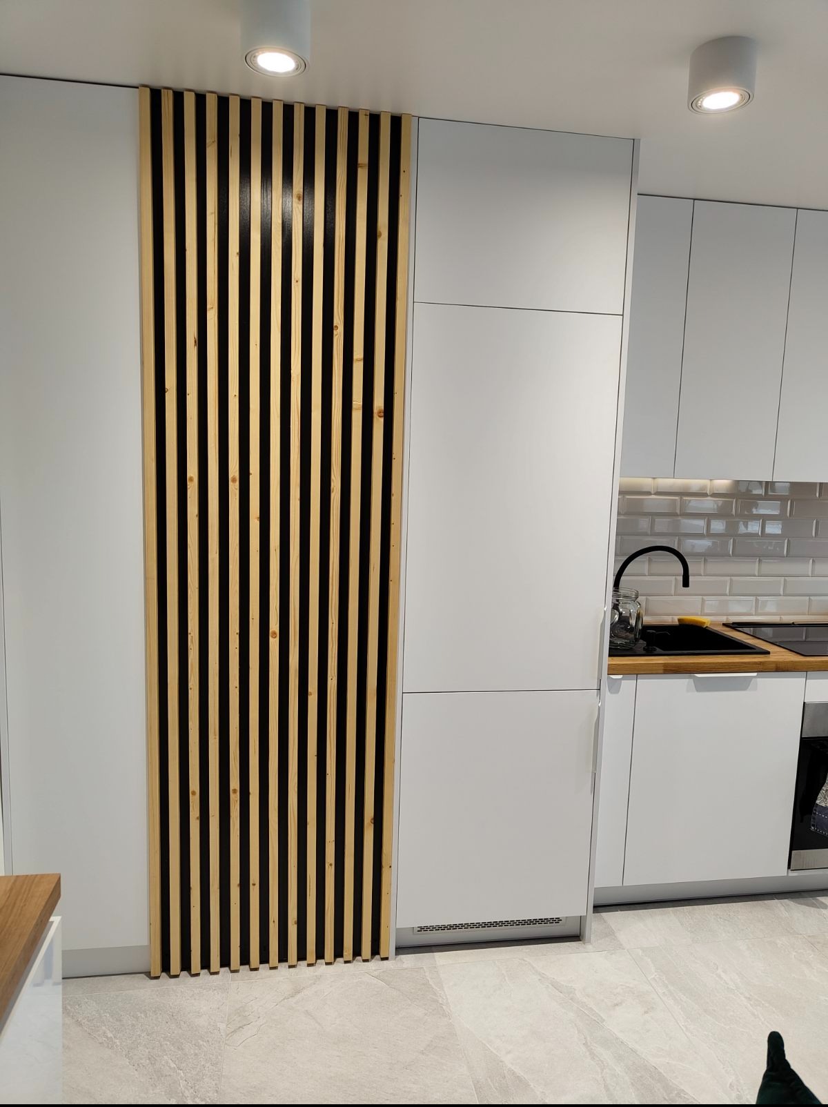 Biała kuchnia z drewnianą ścianką działową z pionowych listew, minimalistyczny design, jasne oświetlenie, szare płytki podłogowe. Nowoczesne wykończenie.