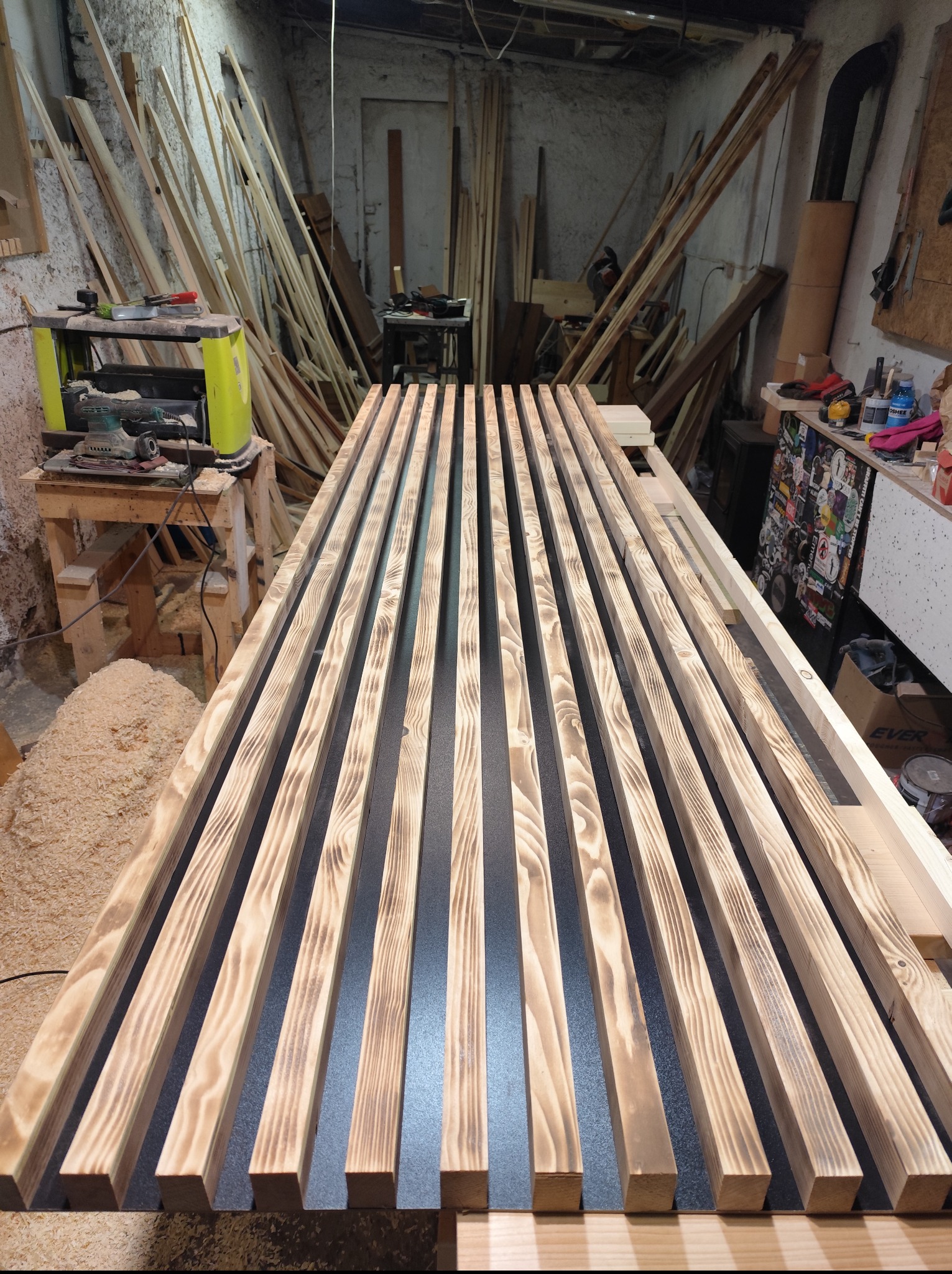 Równoległe, opalane deski z widocznym usłojeniem, ułożone na stole w warsztacie, z narzędziami i stosem drewna w tle. Drewno przygotowane do montażu.