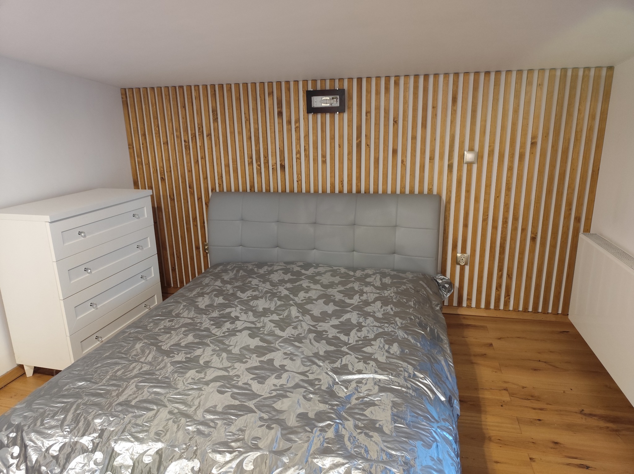 Sypialnia z łóżkiem i drewnianą ścianą z pionowych listew. Biała komoda po lewej. Podłoga z paneli. Na ścianie zegar i włącznik światła.