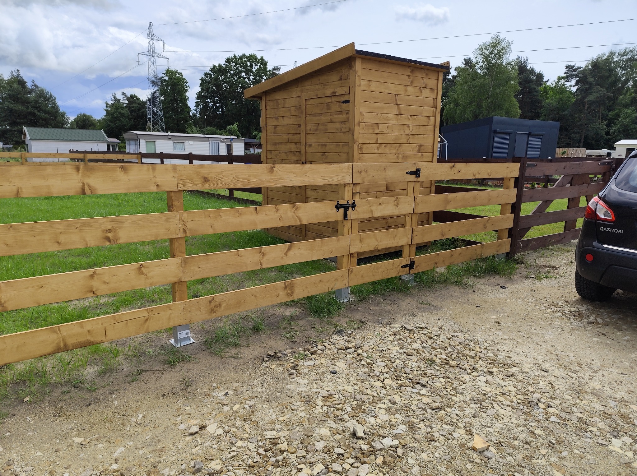 Nowe, jasne ogrodzenie z drewnianą bramką i zamykaną budką w tle, na metalowych wspornikach. Fragment auta po prawej. Trawa i kamienista nawierzchnia.