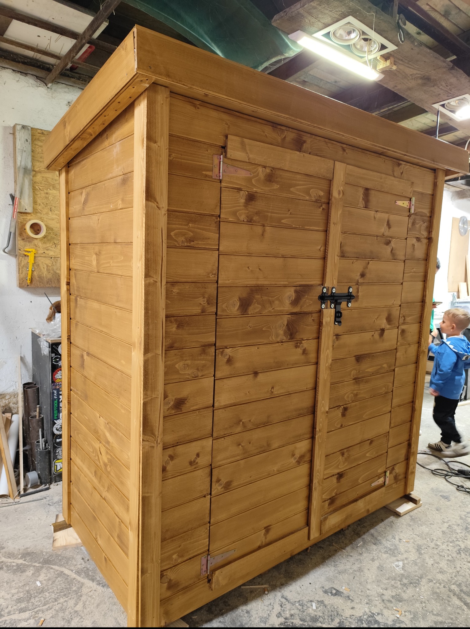 Drewniana, nowa, dwudrzwiowa szopa ogrodowa z metalowym zamknięciem, świeżo po impregnacji, stoi w warsztacie na podkładkach. W tle narzędzia i dziecko.