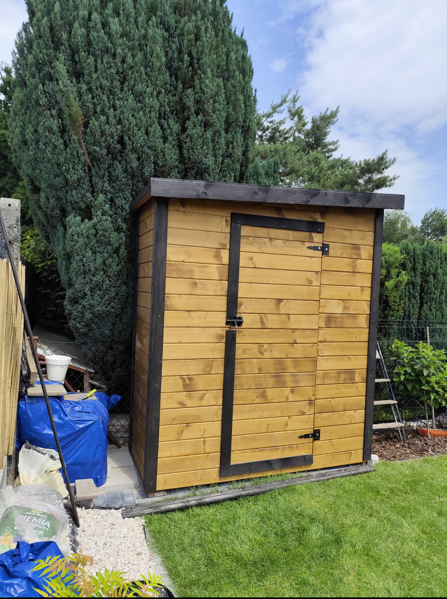 Drewniana, impregnowana szopa ogrodowa z czarnymi elementami wykończeniowymi, umieszczona na trawniku w ogrodzie, na tle zieleni drzew i krzewów.