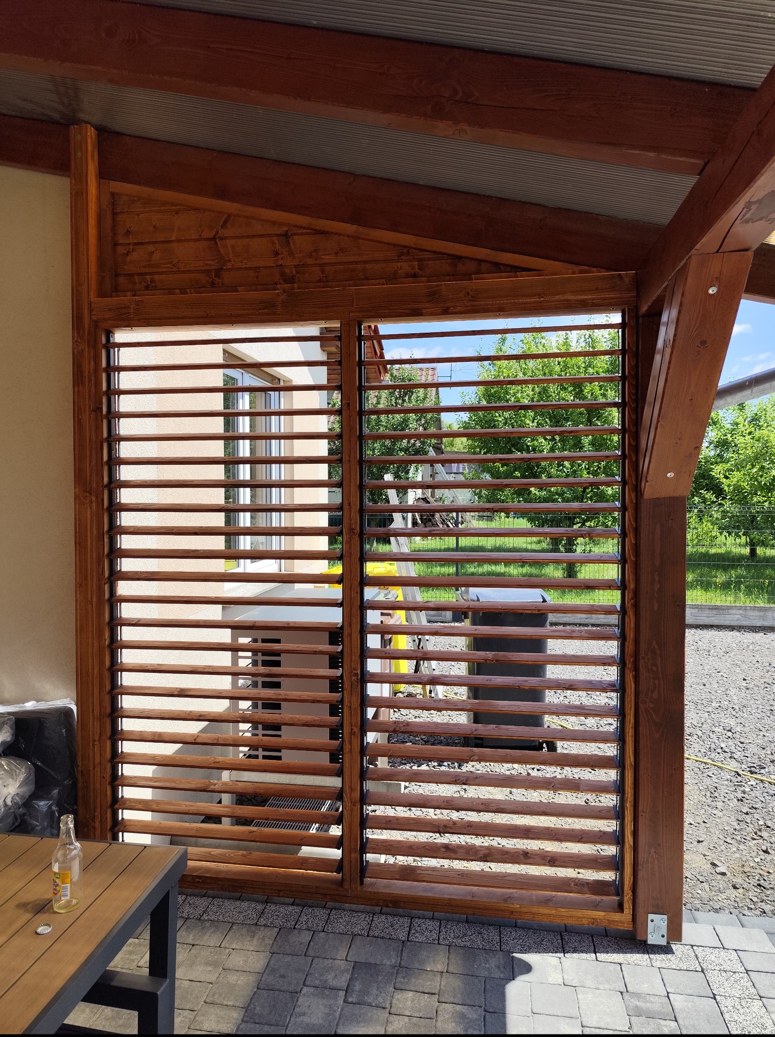 Drewniane żaluzje w brązowej ramie, tworzące ażurową ścianę altany. Widoczny fragment stołu z butelką. W tle zieleń ogrodu i elewacja budynku.