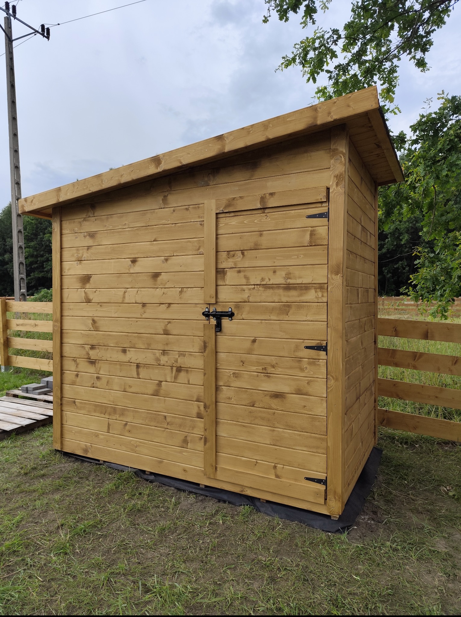 Drewniana, brązowa szopa narzędziowa z czarnymi okuciami, ustawiona na czarnej macie na trawie, z fragmentem drewnianego ogrodzenia w tle.