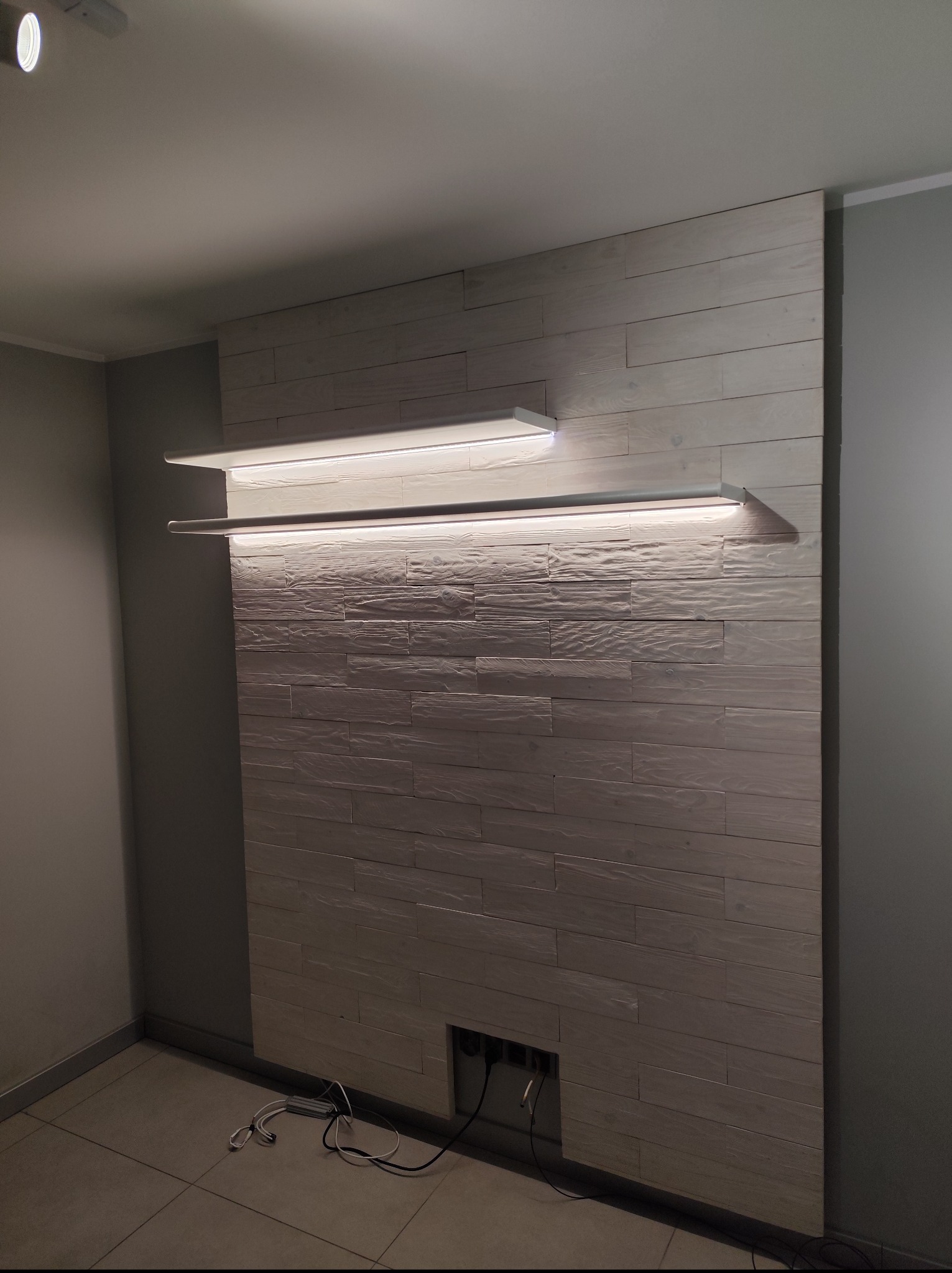 Ściana z paneli drewnianych z oświetleniem LED na półkach. Widoczne kable i otwór w dolnej części ściany. Nowoczesny design z teksturą drewna.