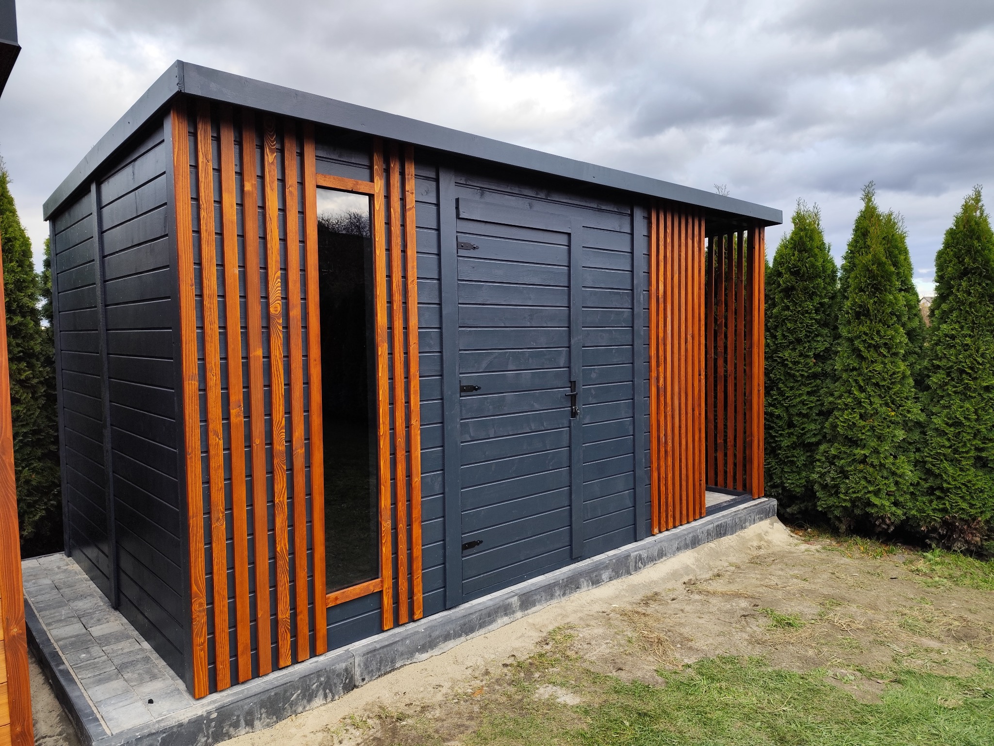 Nowoczesna, grafitowa altana z drewnianymi, pionowymi listwami w kolorze miodowym, z oknem i drzwiami, na tle pochmurnego nieba i zielonych tuj.
