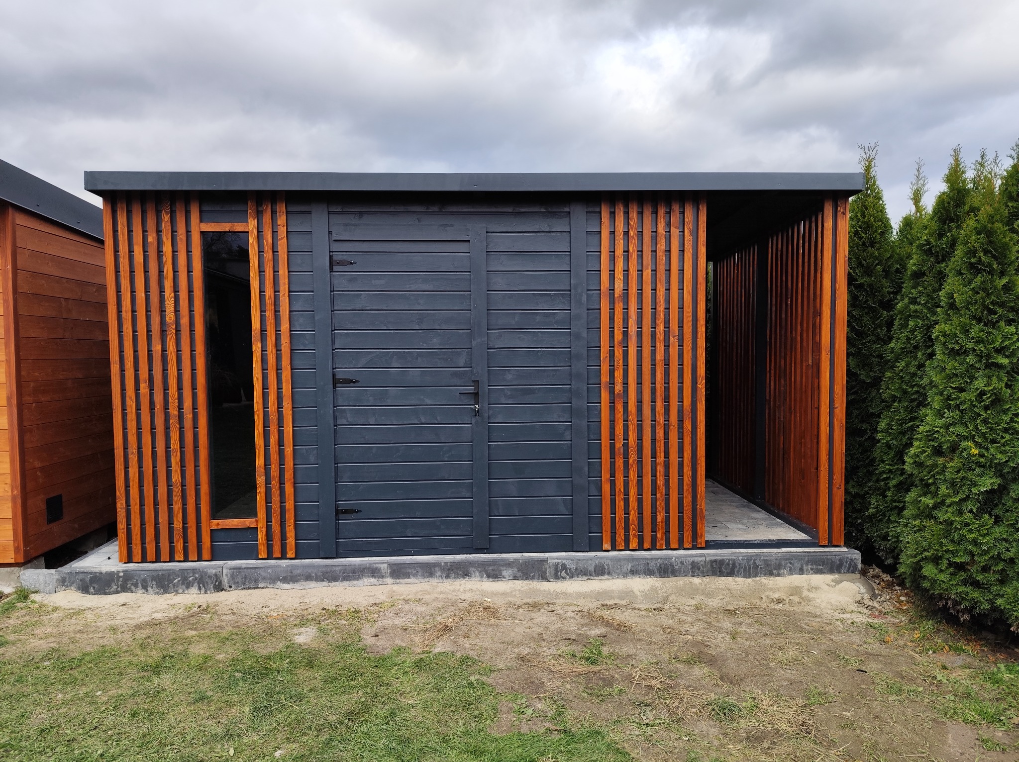 Nowoczesna, grafitowa altana z drewnianymi lamelami w odcieniu miodowym, z widocznymi drzwiami i fragmentem sąsiedniej konstrukcji. Całość na betonowej podmurówce.