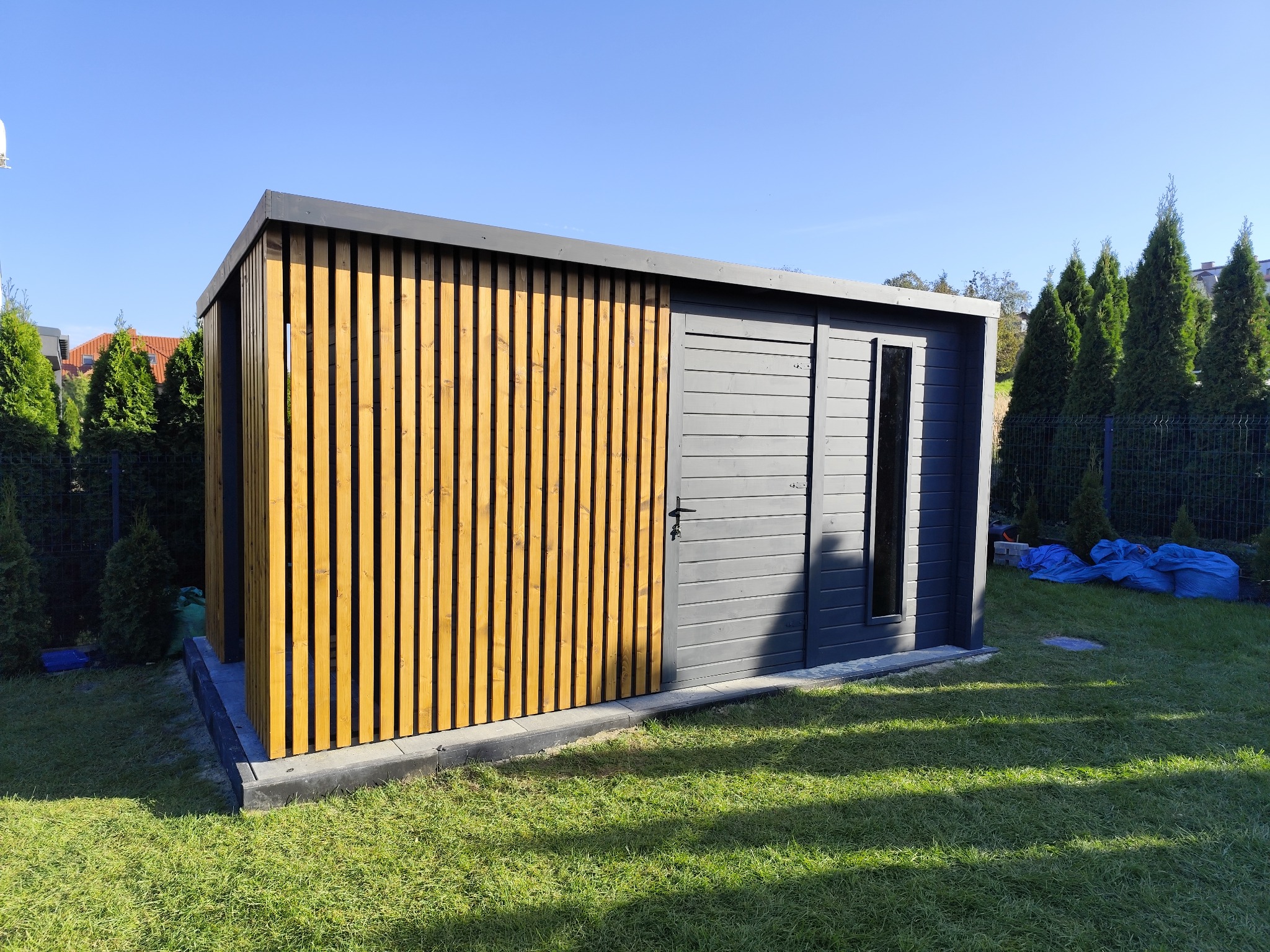 Nowoczesna, szara drewutnia z elementami z jasnego drewna, zlokalizowana w ogrodzie na tle zielonej trawy i iglaków, w słoneczny dzień.