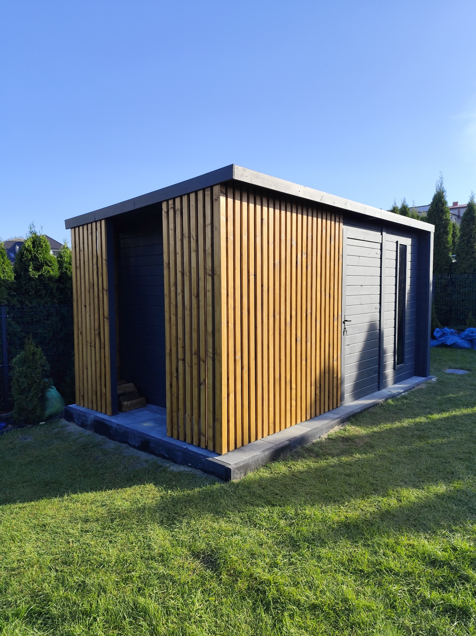 Nowoczesna, szara altana z drewnianymi lamelami w odcieniu miodowym, z drzwiami i oknem, na betonowej podstawie, otoczona zielonym trawnikiem i tujami w słoneczny dzień.