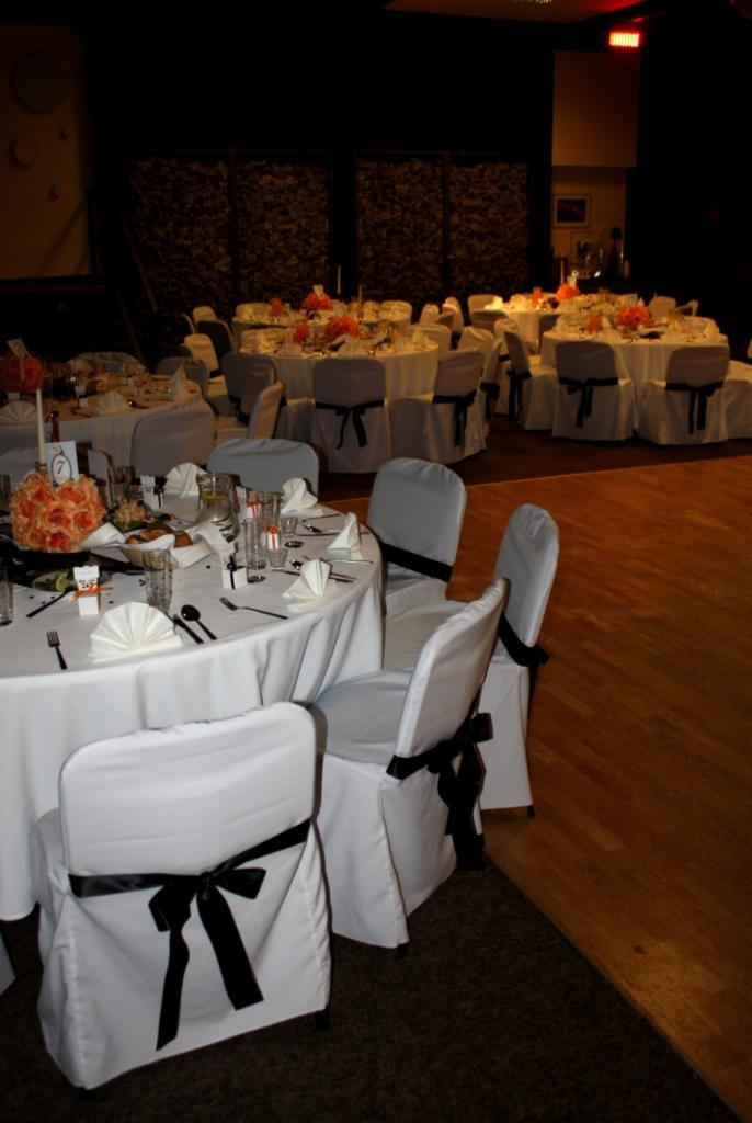 Uroczysta sala bankietowa z okrągłymi stołami przykrytymi białymi obrusami, każdy stół z pomarańczowymi kwiatowymi dekoracjami i nakryciami, krzesła z białymi pokrowcami i czarnymi kokardami...