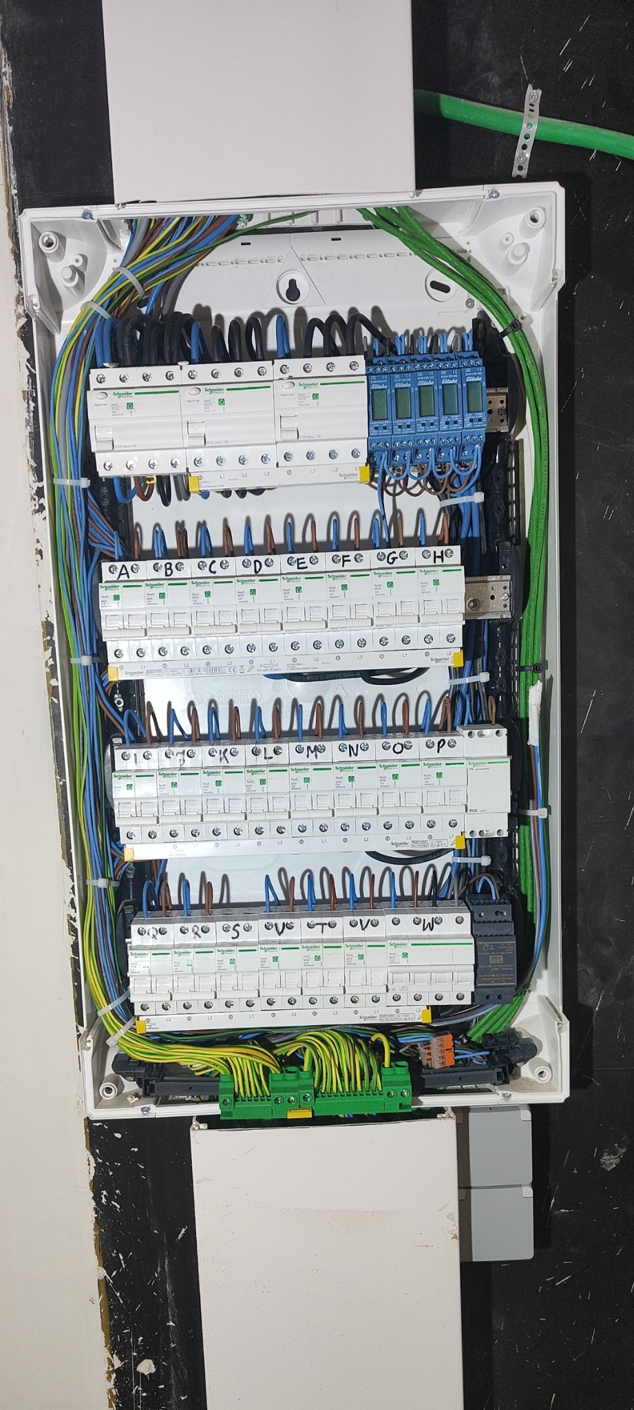 Rozdzielnica elektryczna z bezpiecznikami i przewodami różnych kolorów, zamontowana w białej obudowie na ścianie z widocznymi śladami po malowaniu.