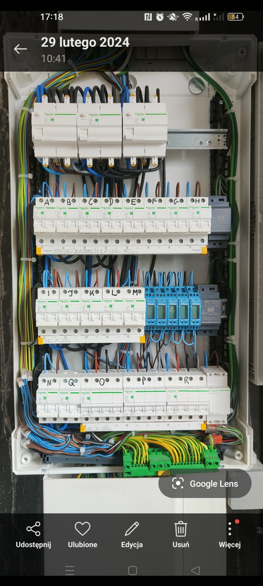 Wnętrze skrzynki elektrycznej z zamontowanymi bezpiecznikami, modułami i okablowaniem oznaczonym literami od A do R, w kolorach niebieskim, czarnym, brązowym i zielono-żółtym.