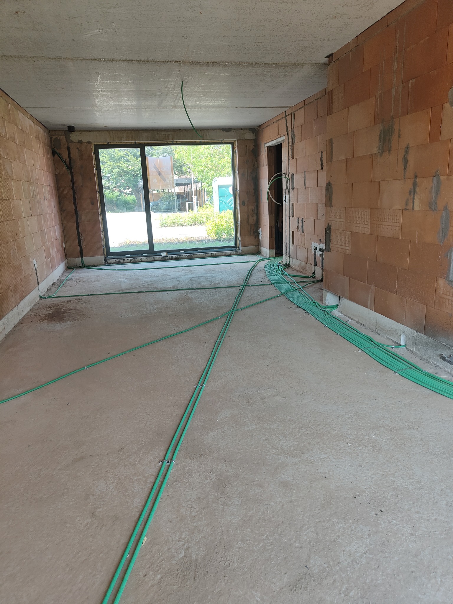 Instalacja elektryczna w surowym wnętrzu: zielone kable ułożone na podłodze, prowadzące do ścian z cegły. Widok na okno i zieleń na zewnątrz.