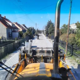 Widok z kabiny żółtej maszyny budowlanej na ulicę w trakcie układania kostki brukowej; w oddali pracownicy, po bokach domy i ogrodzenia.