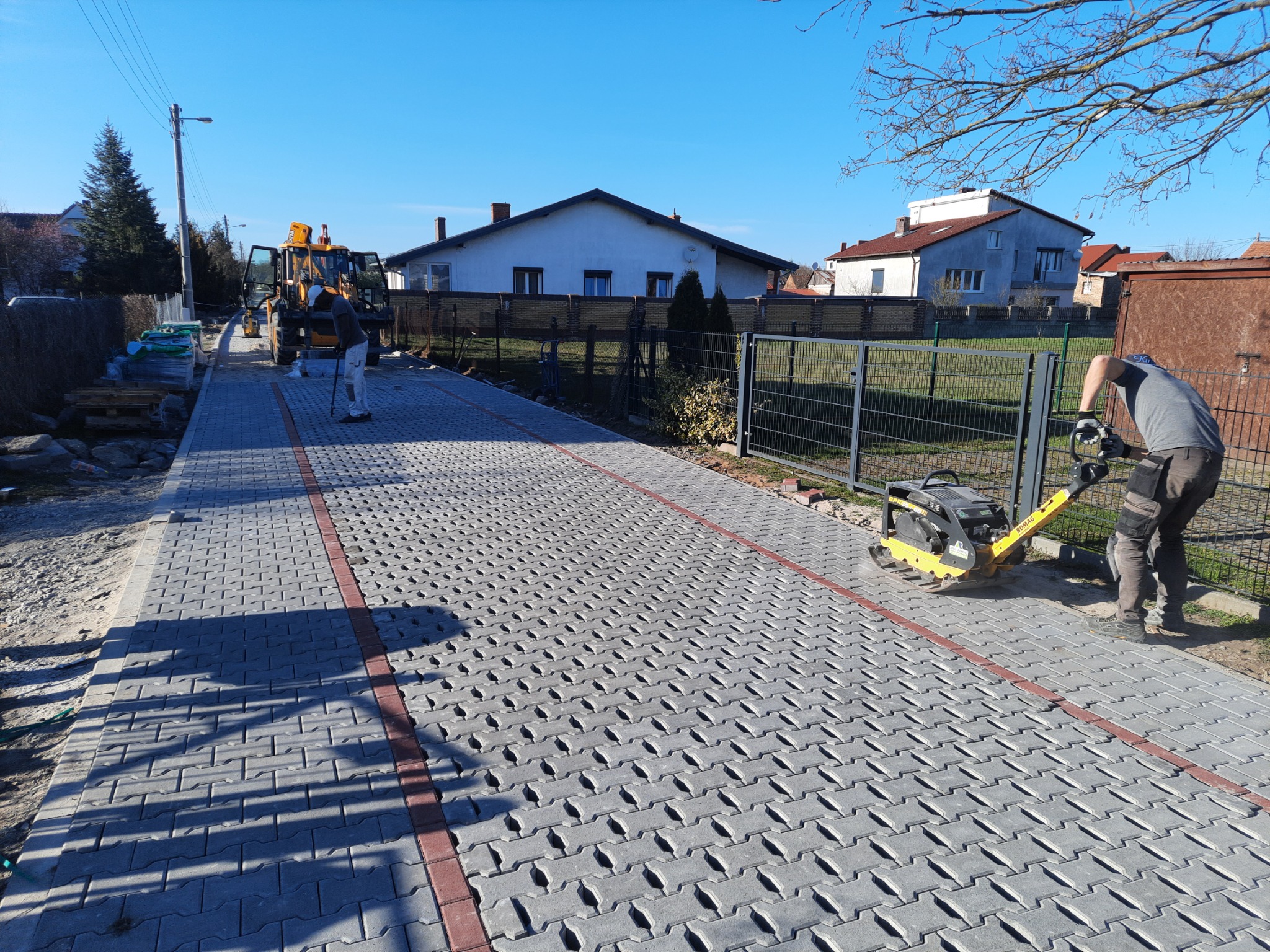 Prace brukarskie w toku: zagęszczarka wibrująca ubija ułożoną szarą kostkę brukową w wzór jodełki, z czerwoną obwódką; w tle koparko-ładowarka i pracownicy.