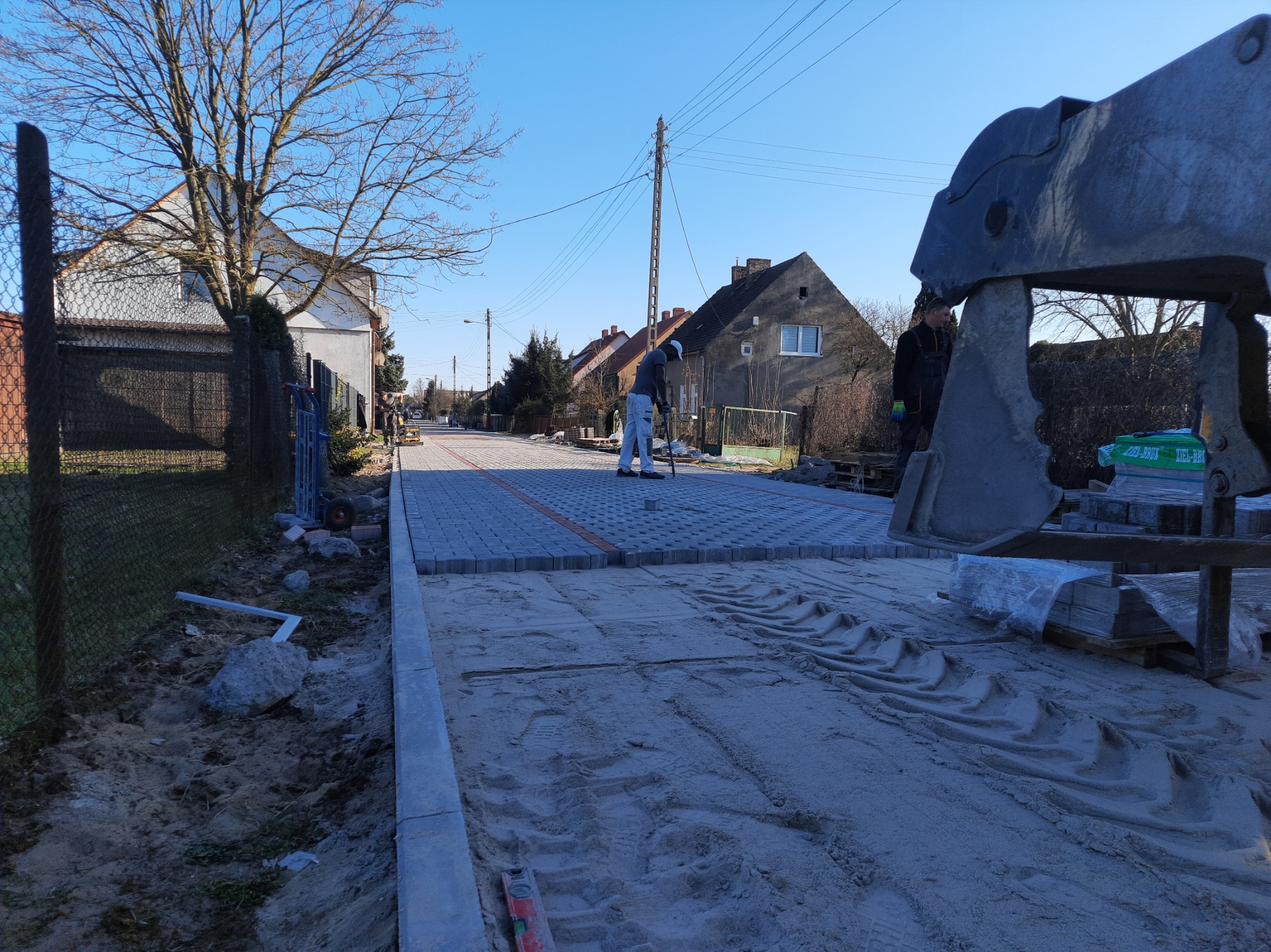 Pracownik wyrównuje podłoże pod kostkę brukową na ulicy osiedlowej, widoczny ciężki sprzęt budowlany i częściowo ułożona nawierzchnia z szarych kostek, w tle zabudowa jednorodzinna i słupy...