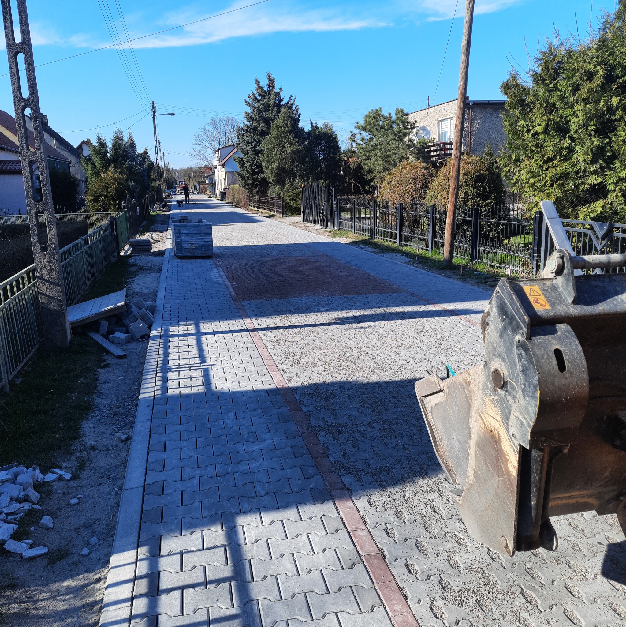 Układanie kostki brukowej na ulicy w trakcie realizacji, widoczny fragment koparki, ułożona kostka w szarym i czerwonym kolorze, niezabudowany fragment drogi z piaskiem.