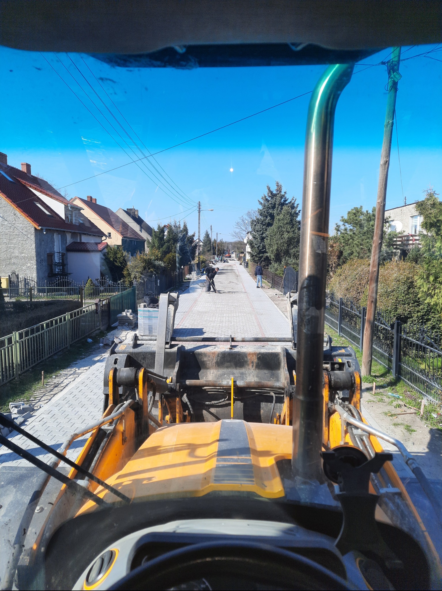 Widok z kabiny żółtej maszyny budowlanej na ulicę w trakcie układania kostki brukowej; w oddali pracownicy, po bokach domy i ogrodzenia.