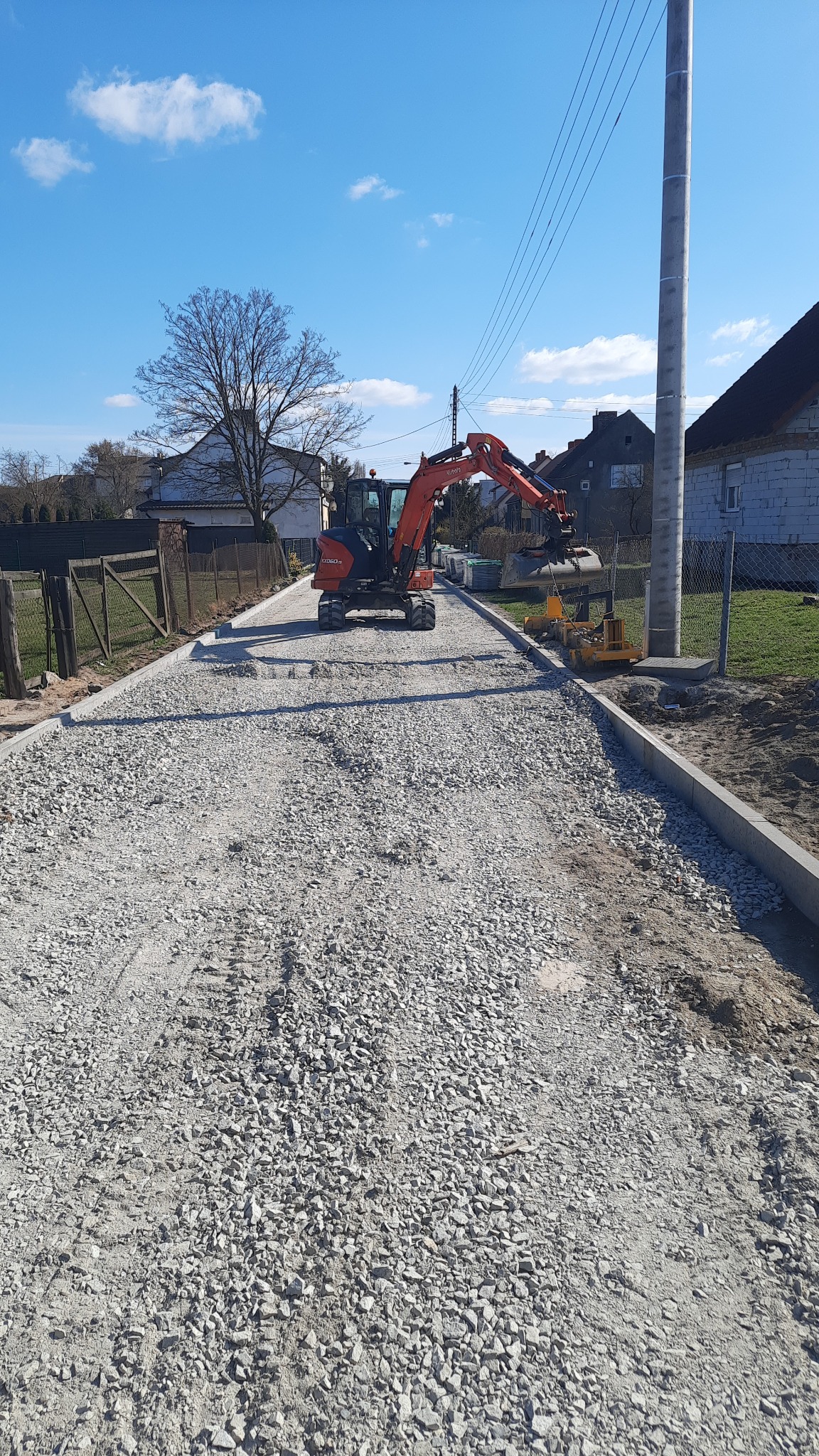 Pomarańczowa minikoparka Kubota na drodze z tłucznia, obok ułożone krawężniki, w tle domy jednorodzinne i słup energetyczny.