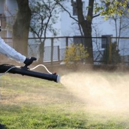 RATATUJ 24 RYSZARD RESZCZYŃSKI - Pracownik w kombinezonie ochronnym i masce rozpyla preparat z opryskiwacza Stihl na trawniku, widoczne drzewa i budynek w tle.
