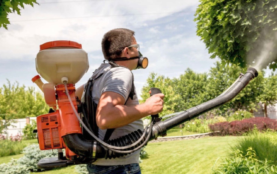 Mężczyzna w masce ochronnej, z opryskiwaczem plecakowym, aplikuje preparat na roślinność w ogrodzie, widoczne rozpylanie substancji.