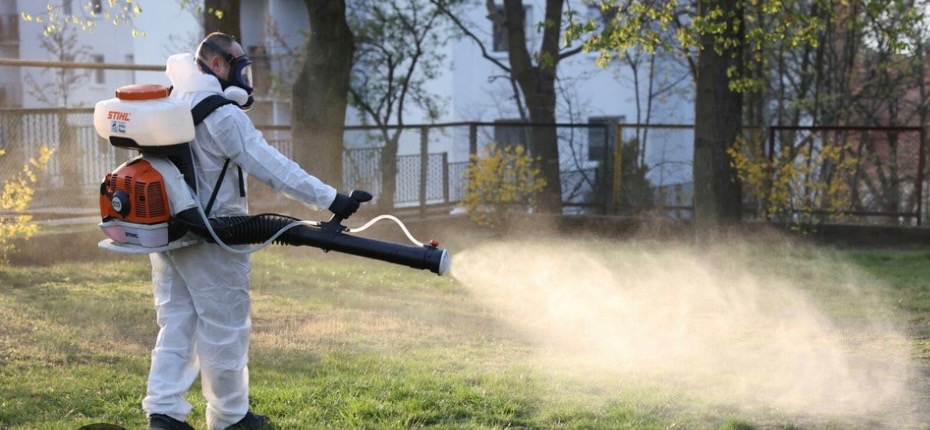 Pracownik w kombinezonie ochronnym i masce rozpyla preparat z opryskiwacza Stihl na trawniku, widoczne drzewa i budynek w tle.